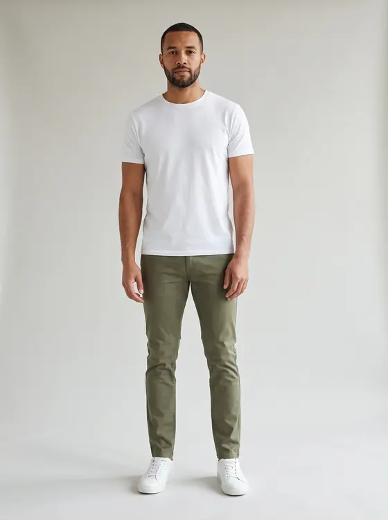 Man with a short beard in olive chinos, a white t-shirt and white sneakers, full length, grey paper backdrop