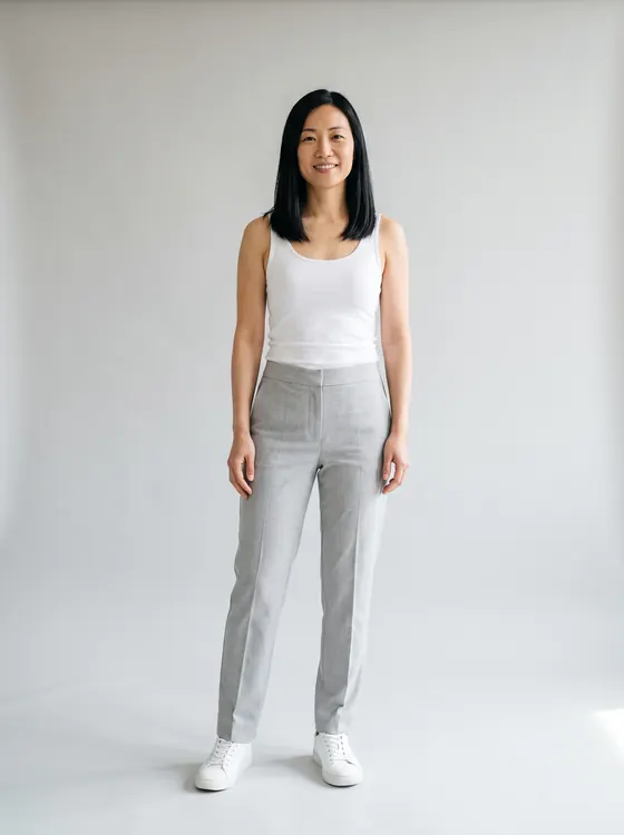 Woman with straight black hair in light grey tailored trousers, a white tank top and white sneakers, full length
